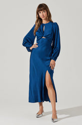 A woman is wearing a long, royal blue satin dress with long, puffed sleeves gathered at the wrists. The dress features a small keyhole opening at the neckline and a cutout detail at the waist. It has a high slit up one side, revealing her leg and silver high-heeled shoes.
